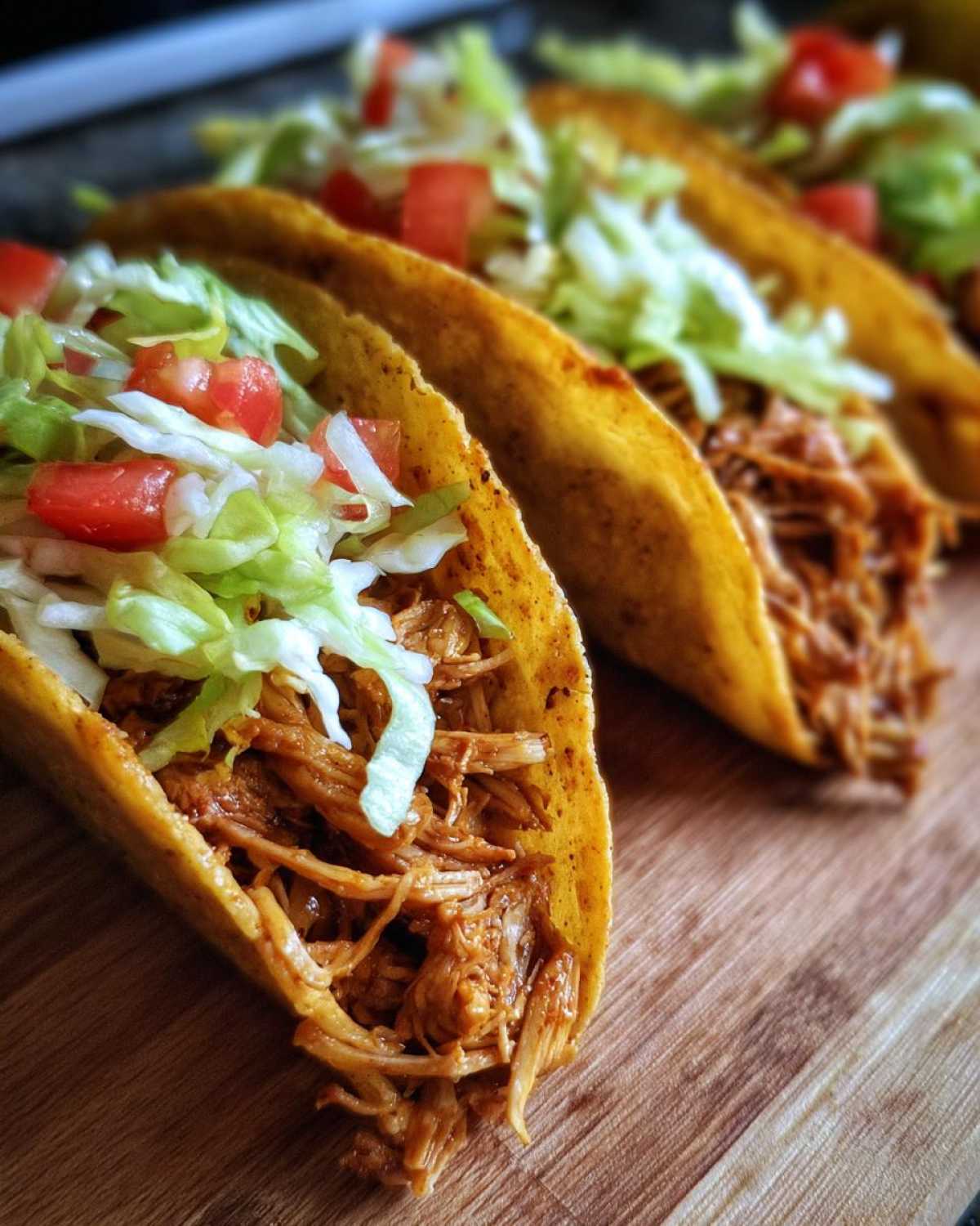 Close-up of crispy mini chicken tacos filled with shredded chicken, lettuce, and diced tomatoes on a wooden board.