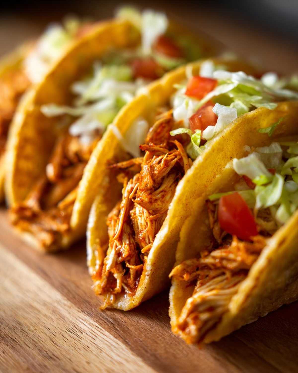 Close-up of crispy mini chicken tacos filled with shredded chicken, lettuce, and tomatoes on a wooden board.