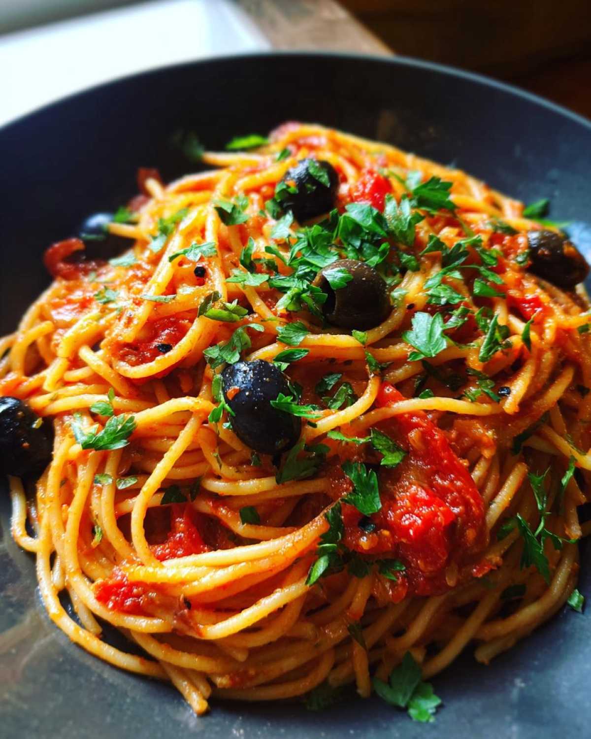 Close-up of one pan spicy pasta in a bowl, topped with tomato sauce, olives, and fresh parsley.