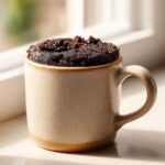 Close-up of a freshly baked Oreo mug cake in a beige mug, topped with crushed Oreos.