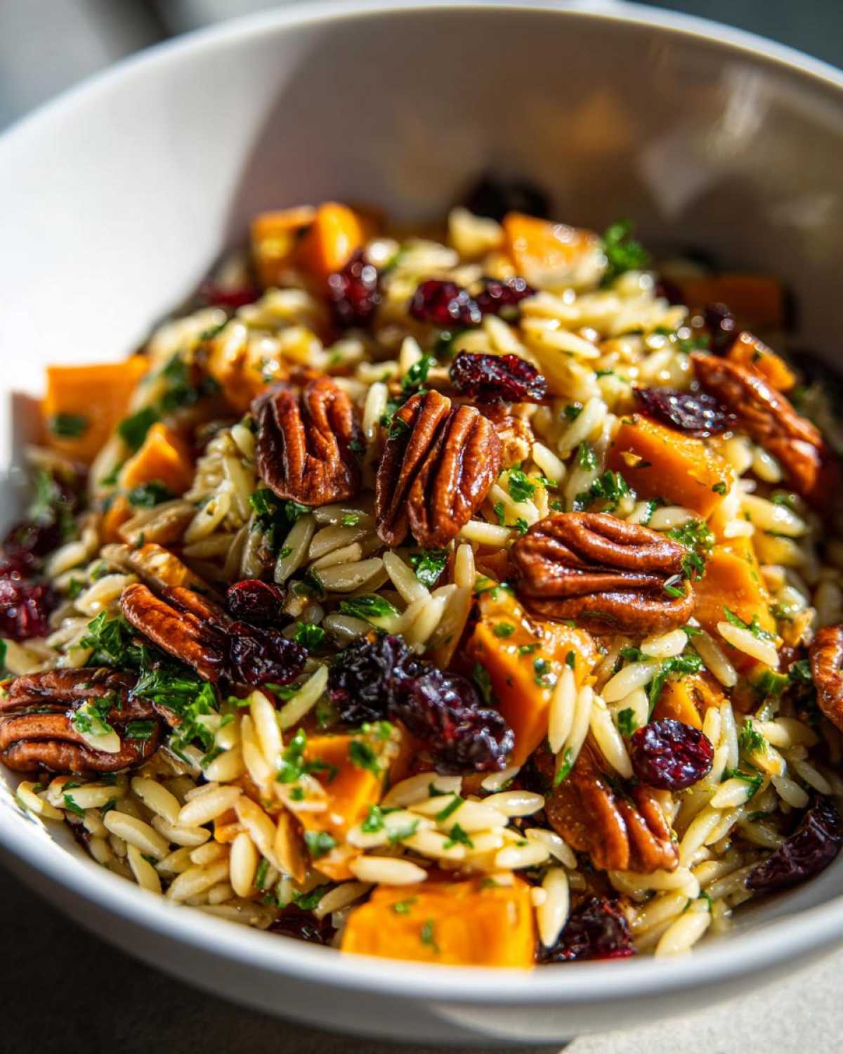 Close-up of a bowl of orzo sweet potato salad with pecans, cranberries, and fresh herbs.