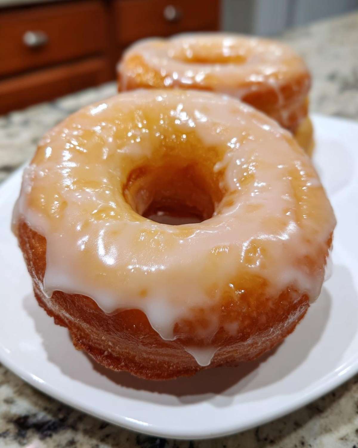 Two donuts on a white plate, featuring a shiny donut glaze dripping down the sides.