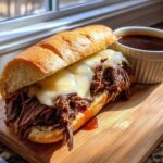 A cheesy slow cooker french dip sandwich on a wooden board with au jus.