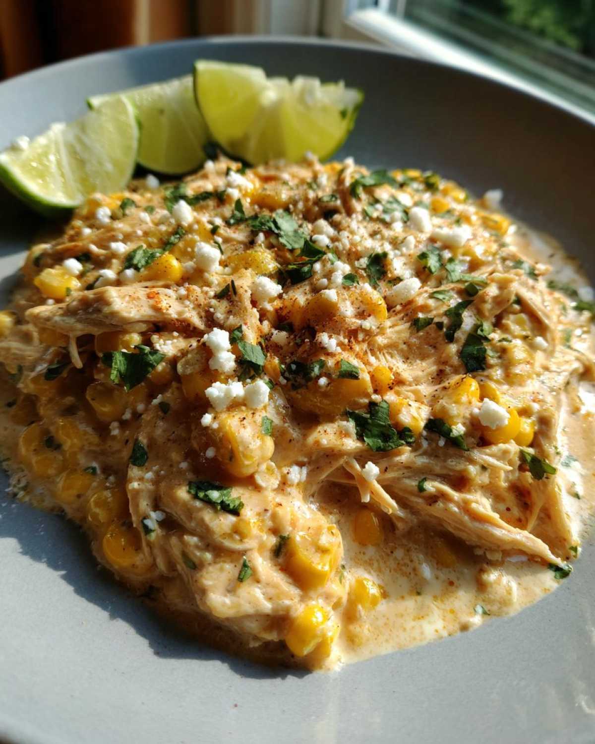 A plate of creamy slow cooker street corn chicken, garnished with cilantro, cotija cheese, and lime wedges.