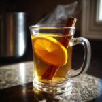 A steaming glass mug of apple cider with a slice of orange and cinnamon stick.