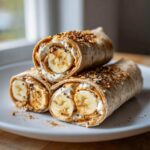 Stack of three high protein banana roll-ups with peanut butter, yogurt, and seeds on a white plate.