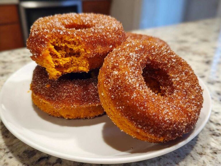 A stack of three golden brown baked pumpkin donuts coated heavily in cinnamon sugar, one with a bite taken out.