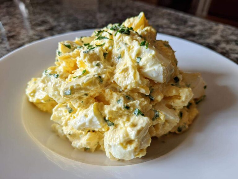 A mound of creamy, chunky egg salad garnished with fresh green herbs on a white plate.