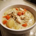 A close-up of creamy slow cooker chicken stew with shredded chicken, carrots, and potatoes, topped with herbs.