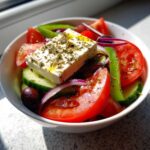 Close-up of a vibrant greek salad featuring a large block of feta cheese drizzled with olive oil and herbs.