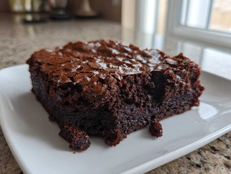 A close-up of a rich, dark chocolate brownies square with a classic shiny, crackly top crust on a white plate.