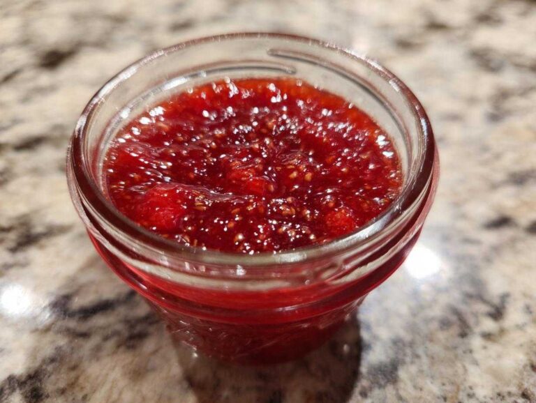 Close-up view of bright red strawberry freezer jam filled in a small glass jar sitting on a granite countertop.