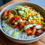 A close-up of an amazing shrimp bowl featuring grilled shrimp over white rice, topped with corn salsa, guacamole, and a creamy green sauce.
