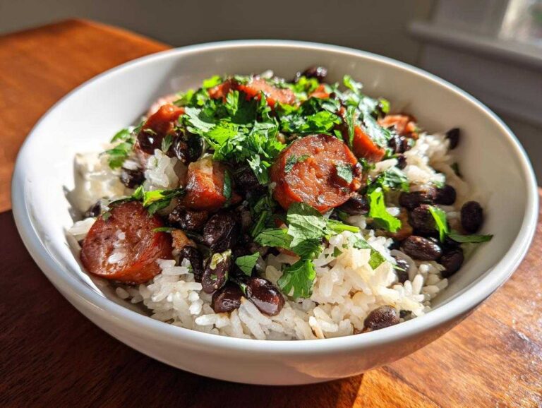Close-up of a white bowl filled with white rice, black beans, sliced smoked sausage, and fresh cilantro, representing a delicious beans and rice dish.