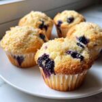 Five freshly baked blueberry protein muffins with a sugary crumble topping arranged on a white plate.