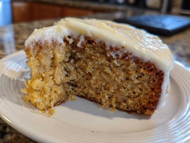 Close-up of a moist slice of applesauce cake topped with thick, creamy white frosting on a white plate.
