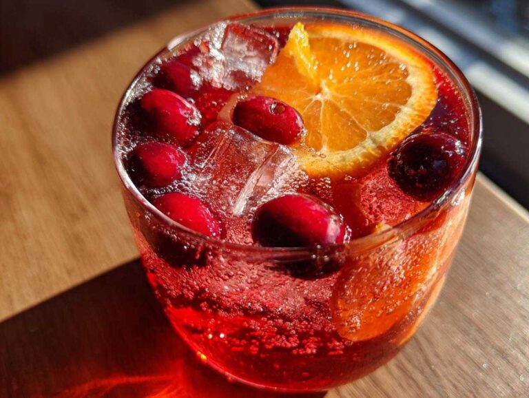 Close-up of a glass filled with bubbling, red non-alcoholic Christmas punch, garnished with fresh cranberries and an orange slice.