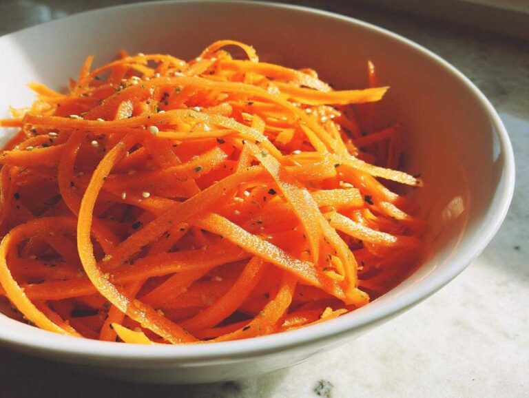 Close-up of vibrant orange raw carrot salad, julienned and sprinkled with sesame seeds and herbs, in a white bowl.