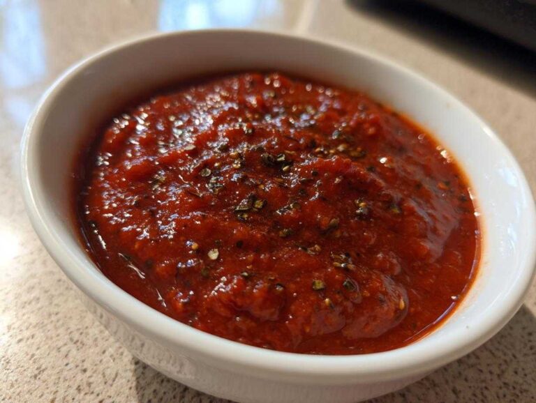 Close-up of rich, vibrant red pizza sauce seasoned with herbs, served in a small white bowl.
