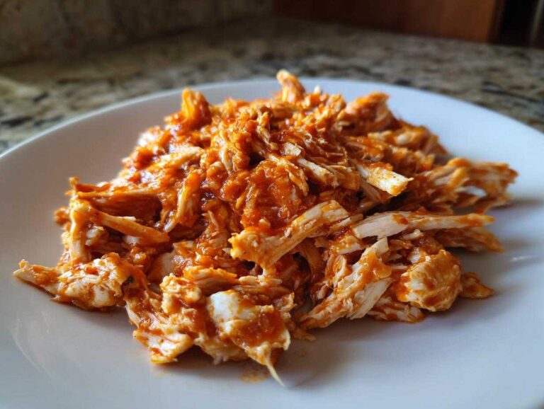 Close-up of savory shredded chicken tinga piled on a white plate, coated in a rich, orange-red sauce.