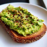Close-up of creamy, mashed avocado toast topped generously with red pepper flakes and black pepper on toasted seeded bread.
