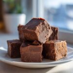 A stack of rich, dark chocolate fudge squares resting on a white plate near a window.