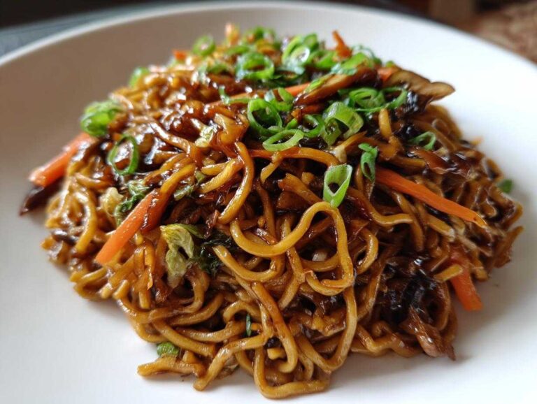A close-up shot of a serving of glossy, dark sauced fried noodles topped with bright green scallions and julienned carrots.