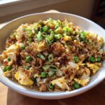 A close-up of a white bowl filled with amazing chicken fried rice, featuring scrambled egg, peas, and fresh green onions.