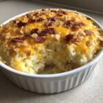 A scoop has been taken out of a rich potato casserole topped with melted cheddar cheese, bacon bits, and crispy onions.