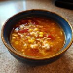 Close-up of a bowl of rich, orange broth maryland crab soup filled with shredded crab meat, bright yellow corn, and diced tomatoes.