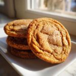 A stack of three golden brown butter sugar cookies, sparkling with granulated sugar, sitting on a white plate near a window.