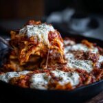 A serving of rich, cheesy skillet lasagna being lifted out of a dark pan, showing a dramatic cheese pull.