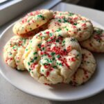 A close-up of soft, round christmas sprinkle sugar cookies topped with red and green nonpareils, served on a white plate.