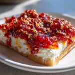 A close-up of a serving of pepper jelly cheese dip, featuring a thick layer of cream cheese topped with vibrant red pepper jelly and sesame seeds.