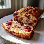 A close-up of a freshly baked cranberry walnut bread loaf with one slice cut, showing bright red cranberries throughout the crumb.