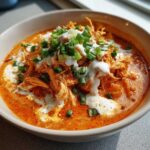 Close-up of a bowl of rich, orange buffalo chicken chili topped with shredded chicken, sour cream swirls, and fresh green onions.