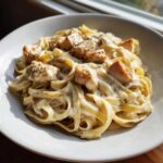 A close-up of creamy chicken fettuccine alfredo pasta topped with seasoned chicken chunks in a white bowl.