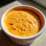 Close-up of creamy, orange cottage cheese queso dip speckled with red peppers and herbs in a white bowl.