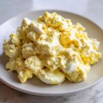 A close-up of a generous serving of creamy, yellow egg salad recipe speckled with black pepper on a light gray plate.