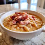 Close-up of a white bowl filled with creamy navy bean soup, topped generously with shredded ham and crispy bacon pieces.