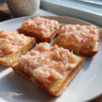 Four pieces of crispy shrimp toast topped with a creamy pink shrimp mixture, served on a white plate in bright sunlight.