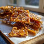 Close-up of shiny, broken pieces of homemade peanut brittle recipe stacked on parchment paper.