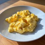A serving of bright yellow, fluffy scrambled eggs piled on a white plate, highlighted by sunlight.