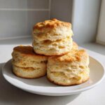 A stack of three perfectly baked, tall, flaky scones with golden tops resting on a white plate.