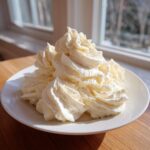 A mound of perfectly piped, fluffy vanilla buttercream frosting on a white plate near a window.