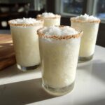 Four frosty glasses of white christmas margarita topped with shaved ice and a toasted coconut rim.