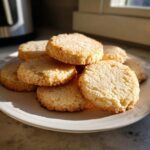 A stack of freshly baked, golden brown sugar cookies piled on a white plate, catching bright sunlight.
