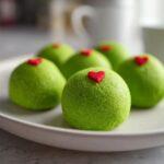 Close-up of several bright green Grinch Oreo Balls, each topped with a tiny red heart sprinkle, resting on a white plate.