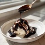A spoonful of rich chocolate syrup is drizzled over a scoop of vanilla ice cream in a white bowl.