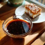 Close-up of a small glass bowl filled with dark, glossy homemade teriyaki sauce, with a piece of glazed food blurred in the background.